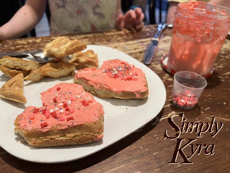 Image shows a closeup of one of the plates with two hearts coated in pink cream cheese and sprinkles. The edge pieces aren't decorated. Beside it sits the cream cheese and container of extra sprinkles while Zoey is blurred in the back waiting to eat it. 